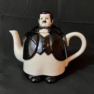 Puede incluir: Una tetera novedosa con forma de hombre con esmoquin. La tetera es principalmente blanca con detalles negros, incluido el esmoquin, la pajarita y los pies. El asa y el pico son blancos.