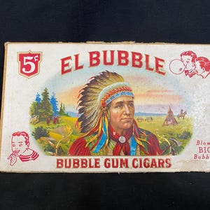 May include: A vintage box of El Bubble Bubble Gum Cigars. The box is white with red and black lettering. The front of the box features a Native American man wearing a headdress and a red, white, and blue shirt. The box also features a cartoon image of a boy blowing a bubble. The text on the box reads "5¢ El Bubble Blows Big Bubbles Bubble Gum Cigars".