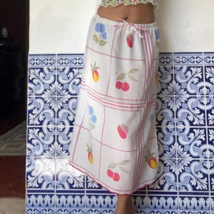 May include: A white skirt with a red grid pattern and a scalloped hem. The skirt is decorated with colorful illustrations of fruits, including cherries, strawberries, and peaches. A pink bow is tied at the waist.