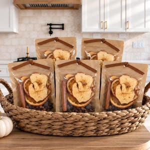 May include: Five clear pouches filled with dried apple slices, orange slices, and cinnamon sticks are arranged in a woven oval basket. A small white pumpkin sits on the wooden surface next to the basket, which contains these aromatic ingredients.