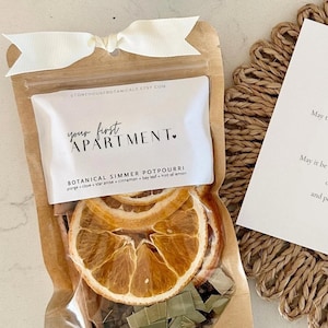 May include: A clear pouch of botanical simmer potpourri with dried orange slices, cinnamon sticks, and bay leaves, topped with a white ribbon. Beside it, a square card reads New Apartment Blessing with a printed poem about new beginnings.