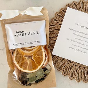 May include: A clear pouch of botanical simmer potpourri with dried orange slices, topped with a white ribbon. Next to it is a square card titled New Apartment Blessing with printed text, resting on a round, braided tan jute coaster.
