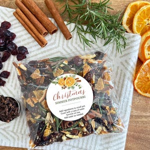 May include: A clear plastic bag filled with Christmas simmer potpourri containing dried orange slices, cranberries, cinnamon sticks, and rosemary. A round white label features the text Christmas SIMMER POTPOURRI and instructions to add 3 cups of water.