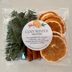May include: A clear plastic bag containing a Cozy Winter simmer potpourri kit. The kit includes dried orange slices, cinnamon sticks, and evergreen sprigs. A round label reads "Cozy Winter" with instructions.