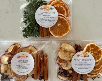 May include: Three clear plastic pouches containing simmer potpourri ingredients. The top pouch labeled COZY WINTER includes pine needles, cinnamon sticks, and dried orange slices. The bottom pouches labeled APPLE PIE and WINTER SPICE contain dried apple slices, cinnamon sticks, star anise, and cloves.