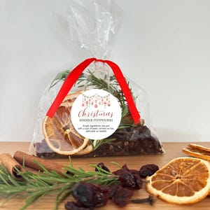 May include: A clear plastic bag filled with dried orange slices, rosemary sprigs, and dark berries, tied with a red ribbon. A circular white label features the text Christmas SIMMER POTPOURRI and instructions to empty ingredients into a pot.