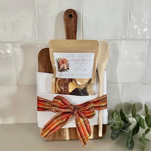 May include: A wooden cutting board wrapped in a white cloth with an orange and red plaid ribbon, holding a Stonehouse Botanicals Autumn Harvest simmer potpourri pouch and a light-colored wooden spoon. The pouch label lists ingredients.