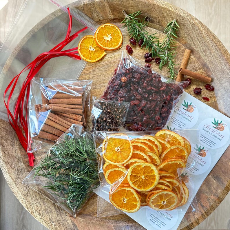 May include: A Christmas simmer potpourri kit containing clear plastic bags filled with dried orange slices, cinnamon sticks, whole cloves, dried cranberries, and fresh rosemary sprigs, accompanied by red ribbon and circular instruction labels.