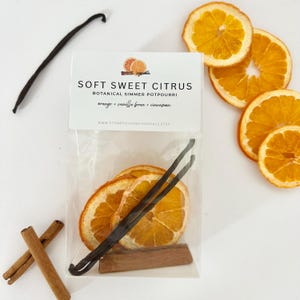 May include: A clear bag containing dried orange slices, vanilla beans, and cinnamon sticks. The bag has a label that reads "Soft Sweet Citrus Botanical Simmer Potpourri." Additional orange slices and a vanilla bean are placed around the bag.