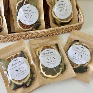 May include: Several kraft paper pouches containing dried citrus slices, bay leaves, and peppercorns. Each pouch features a circular white label with the text: thank you for celebrating with us, S | A, STACY & ALAN, JUNE 27, 2026, and a small leaf icon.