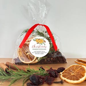 May include: A clear plastic bag filled with dried orange slices, rosemary sprigs, and dark berries, tied with a red ribbon. A circular label reads Christmas SIMMER POTPOURRI and includes instructions to empty ingredients into a pot with 3 cups of water.