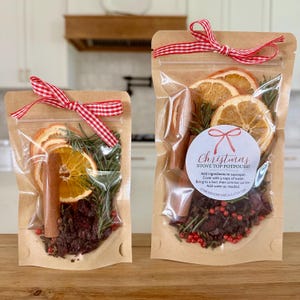 May include: Two clear pouches with kraft paper backing containing stove top potpourri. Each pouch features dried orange slices, cinnamon sticks, rosemary, cranberries, and red peppercorns, tied with a red and white gingham ribbon.