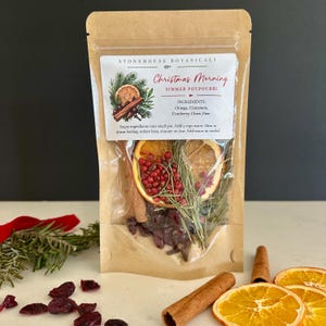 May include: A brown kraft paper pouch containing Christmas Morning simmer potpourri by Stonehouse Botanicals. The label lists ingredients including orange, cinnamon, cranberry, clove, and pine. Dried orange slices, red berries, and pine needles are visible.