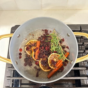 May include: A light gray cooking pot with gold-toned handles containing simmering water, dried orange slices, cinnamon sticks, fresh green rosemary sprigs, and dark red dried cranberries. The pot is positioned on a black stovetop burner grate.