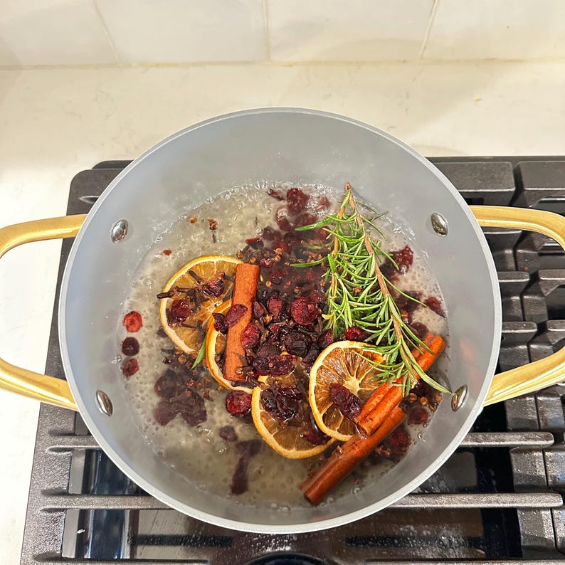 May include: A light gray cooking pot with gold-toned handles containing simmering water, dried orange slices, cinnamon sticks, fresh green rosemary sprigs, and dark red dried cranberries. The pot is positioned on a black stovetop burner grate.