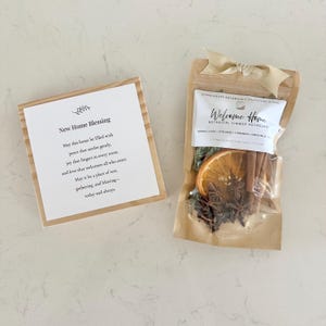 May include: A square wooden card with the text New Home Blessing and a poem, alongside a kraft paper pouch of botanical simmer potpourri topped with a beige ribbon. The pouch label reads Welcome Home and lists ingredients like orange, clove, and cinnamon.