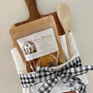 May include: A kraft paper pouch of Stonehouse Botanicals House Blessing simmer potpourri featuring dried orange slices, cinnamon sticks, and bay leaves, tied with a black and white gingham ribbon to a white cloth and a wooden spoon on a wooden board.
