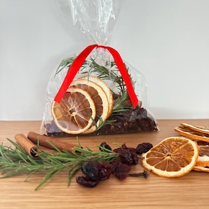 May include: A clear plastic bag tied with a red ribbon containing dried orange slices, rosemary sprigs, and dark berries. Outside the bag are loose cinnamon sticks, fresh rosemary, dried orange slices, and dark berries on a wooden surface.