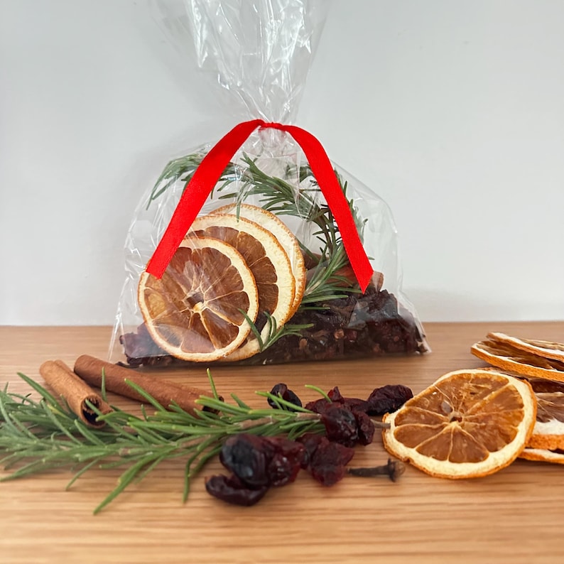 May include: A clear plastic bag tied with a red ribbon containing dried orange slices, rosemary sprigs, and dark berries. Outside the bag are loose cinnamon sticks, fresh rosemary, dried orange slices, and dark berries on a wooden surface.