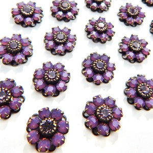 May include: Close-up of several purple flower-shaped embellishments. Each embellishment features a dark metal base with a circular design and is adorned with numerous purple crystals. The center of each flower has a larger crystal.