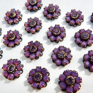 May include: Close-up of several purple flower-shaped embellishments. Each flower has a dark metal base with a ring of purple gems around a central, darker purple gem. The gems have a faceted appearance, catching the light.