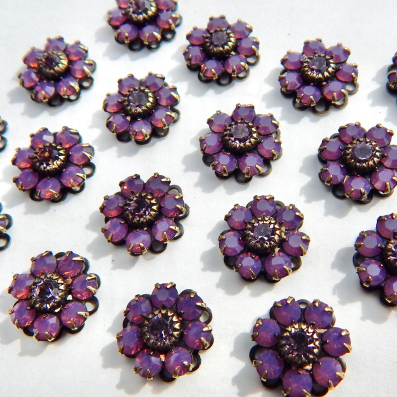 May include: Close-up of several purple flower-shaped embellishments. Each flower has a dark metal base with a ring of purple gems around a central, darker purple gem. The gems have a faceted appearance, catching the light.