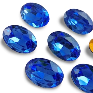 May include: Six oval-shaped, blue, faceted rhinestones. The rhinestones are scattered on a white background.