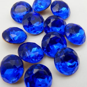 Puede incluir: Una colección de doce gemas de vidrio facetado azul cobalto. Las gemas son redondas y tienen una superficie brillante y reflectante.