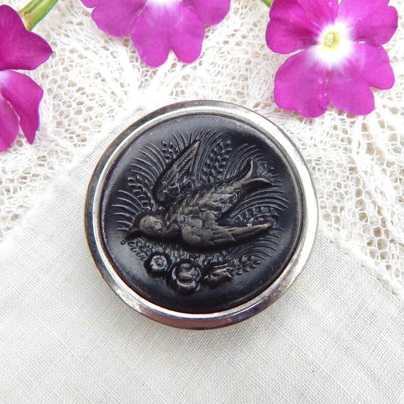 May include: A round, black button with a silver-colored rim. The button features a detailed relief of a bird in flight, surrounded by wheat and floral elements. The button is set against a white lace background, with purple flowers in the corners.
