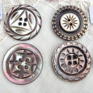 May include: Four decorative buttons with intricate carved designs. The buttons are round, with varying patterns including geometric shapes and floral motifs. The colors range from iridescent pearl to dark gray, with visible holes for sewing. These buttons are ideal for sewing and craft projects.