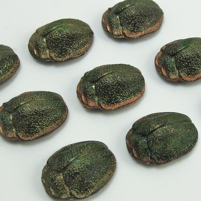 Puede incluir: Siete botones en forma de escarabajo de color verde y cobre con una superficie texturizada.