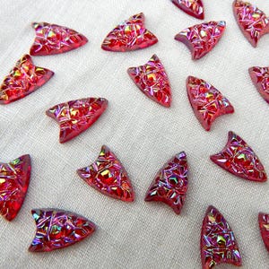 May include: A collection of red, iridescent, arrow-shaped decorative embellishments. Each piece has a faceted surface that catches the light, creating a shimmering effect. The embellishments are scattered on a white fabric surface.
