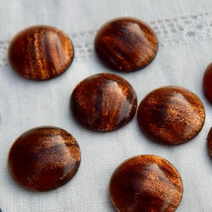 May include: A collection of round, dark brown glass cabochons with a shimmering, metallic copper-colored interior. The cabochons have a domed shape and are arranged on a white, textured surface. These decorative glass pieces could be used for jewelry making or other craft projects.