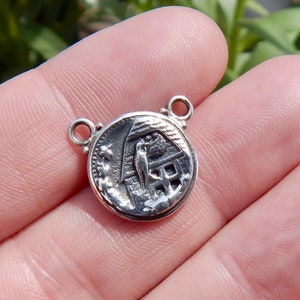May include: A small, round, silver-colored pendant with a detailed engraved design. The pendant features a scene with a bird, a building, and other elements. It has two small loops at the top for attaching to a chain or other jewelry.
