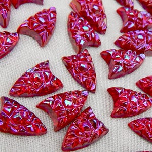 May include: A collection of red, triangular-shaped decorative pieces with a faceted, iridescent surface. The pieces have a textured appearance and are scattered across a neutral background. The overall effect is one of sparkle and shine.