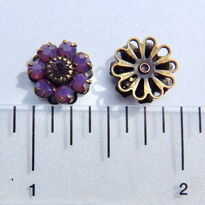 May include: Two antique-style flower-shaped earrings. One earring features purple gemstones arranged around a dark center stone. The other earring has a bronze-colored, openwork design. The earrings are placed on a white surface next to a ruler.