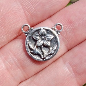 May include: Silver charm with a detailed floral design. The charm is round and has two jump rings for attaching to a necklace or bracelet.