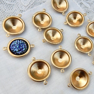 May include: A collection of gold-colored metal jewelry components. The components have a circular, bowl-like shape with small loops for attachment. One component features a blue, iridescent, druzy-like stone. The components are arranged on a white, textured surface.