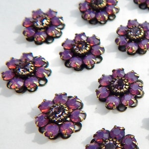 May include: Close-up of several purple flower-shaped embellishments. Each flower has a central, round purple gem surrounded by petal-shaped gems in varying shades of purple. The metalwork is dark with gold accents, creating a vintage look.