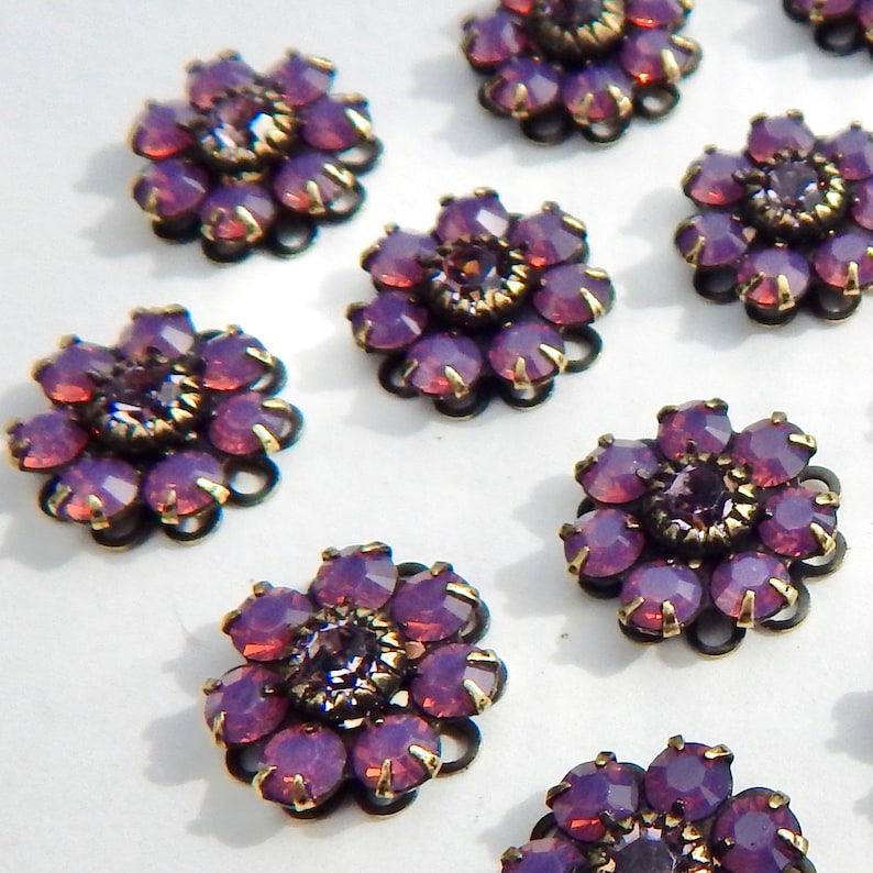 May include: Close-up of several purple flower-shaped embellishments. Each flower has a central, round purple gem surrounded by petal-shaped gems in varying shades of purple. The metalwork is dark with gold accents, creating a vintage look.