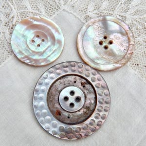 May include: Three iridescent mother-of-pearl buttons. Two are round with four holes, and the third has a textured outer ring and a smaller, central button with four holes. The buttons have a pearlescent sheen with hints of pink, green, and gold.
