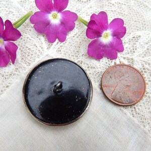 May include: A black button with a silver rim, next to a penny and purple flowers. The button has a loop on the back for sewing. The penny is dated 1979. The flowers are a vibrant purple color.