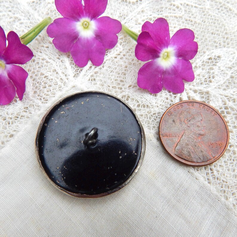 May include: A black button with a silver rim, next to a penny and purple flowers. The button has a loop on the back for sewing. The penny is dated 1979. The flowers are a vibrant purple color.