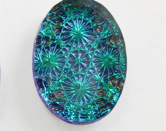 May include: Oval-shaped iridescent glass cabochon with a teal and purple color scheme. The glass has a textured surface with a repeating pattern of starbursts.