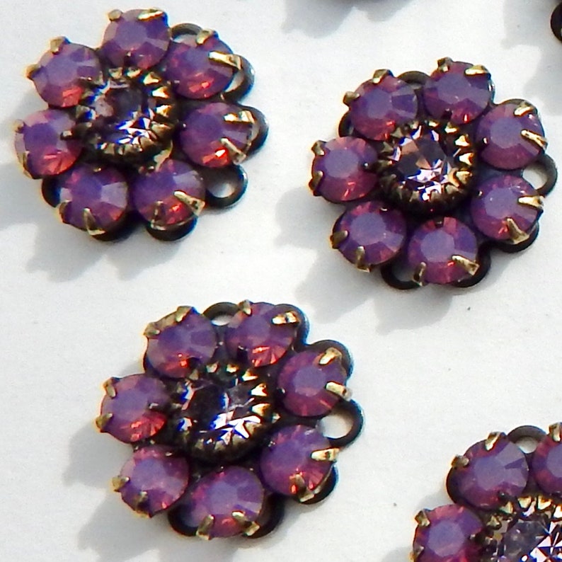 May include: Close-up of several flower-shaped embellishments. Each has a dark metal base with a floral design, and is adorned with purple and pink iridescent gemstones. The center stone is a larger, faceted gem. These could be used for jewelry or craft projects.