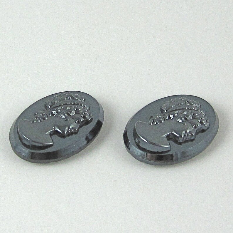 Op de afbeelding: Twee ovale, donkergrijze cameo's. Elke cameo toont een profiel van een vrouw met een lauwerkrans. De cameo's zijn waarschijnlijk gemaakt van gepolijste steen of glas. De achtergrond is effen lichtgrijs.
