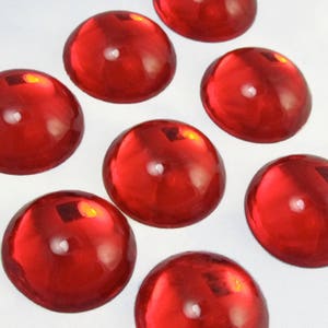 Peut inclure: Huit cabochons ronds en verre rouge brillant. Les cabochons sont disposés en demi-cercle sur une surface blanche.