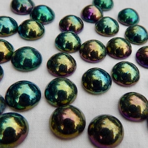 May include: A collection of iridescent, dome-shaped glass beads. The beads display a range of colors, including green, gold, and purple, with a reflective, glossy finish. These decorative glass beads are scattered on a white surface.