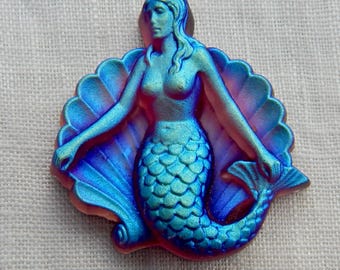 May include: A blue and purple mermaid figurine with a detailed tail and flowing hair, set against a shell-shaped backdrop. The mermaid's arms are outstretched, and the figure has a metallic sheen.