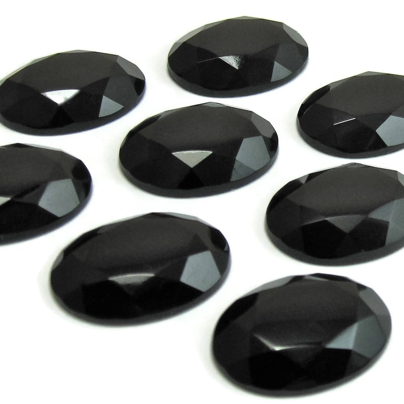 May include: Eight black oval-shaped gemstones with a faceted cut. The gemstones are arranged in a staggered pattern on a white background.